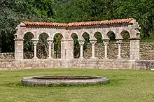 Cloître rénové, mais tout de même en ruines. Devant, au milieu d'une pelouse, un petit bassin. Derrière, des buissons.