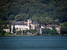 Photographie couleur d'un ensemble de bâtiments monastiques dominés par un clocher et une tour ; photographie prise depuis un bateau, avec le lac au premier plan et la montagne en arrière.