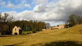 Hauteur boisée descendant en pente douce avec plusieurs bâtiments. Prairie avec une chapelle gothique sans toit