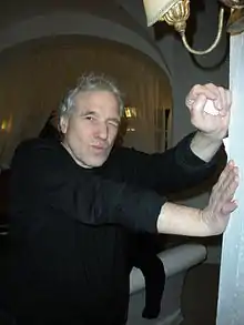 Un homme blanc d'âge mûr, habillé avec un pull-over noir. Il ferme son œil gauche et s'appuie sur un mur