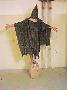 Un homme bras tendus recouvert d'un vêtement, debout sur un carton dans un milieu carcéral.