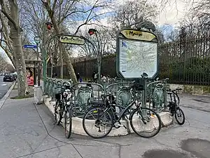 Photographie d'une bouche de métro aux rambardes de laquelle sont accrochés des vélos ; en arrière-plan, une longue perspective animée par une grille sombre aux pointes dorées.