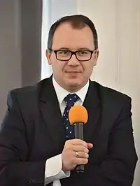 Adam Bodnar en costume noir, cravates et lunettes tenant un micro
