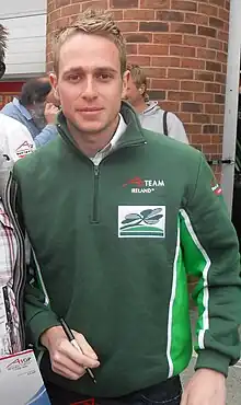 Photographie d'un homme blond, vu de face, souriant, avec un stylo à la main, avec un pull vert foncé.