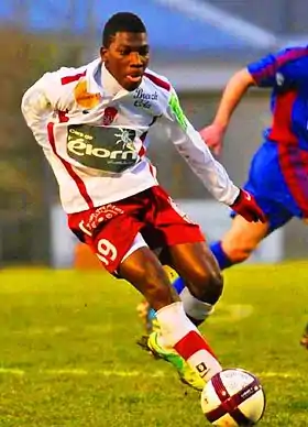 Photographie d'un joueur de football noir au maillot blanc et rouge vu de trois-quarts, courant avec le ballon.