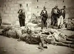 Corps alignés au pied d'un mur, des hommes debout les observent.