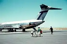 Photographie d'un avion de ligne garé sur le tarmac, avec quatre passagers s'apprêtant à embarquer.