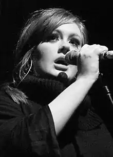 Une jeune femme, Adele, tenant un microphone dans ses mains. La photographie est en noir et blanc