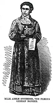Adele Spitzeder est représentée comme une femme à l'allure sévère, en robe ample, avec une grande croix sur la poitrine et une note dans la main gauche.