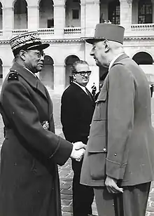 Sur fond de la cour intérieure de l'Hôtel des Invalides, le médecin général Diagne en uniforme avec capote sert la main du Général de Gaulle qui vient de le décorer, à sa gauche Yon Morandat en costume civil lui aussi compagnon de la Libération