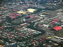 Photographie aérienne d'un complexe sportif comprenant notamment un grand stade de football et plusieurs cours de tennis. L'ensemble est entouré d'habitations rudimentaires.