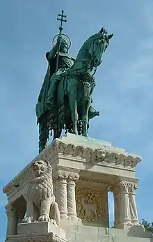 Statue en bronze d'un cavalier couronné tenant un goupillon. La sculpture se trouve au sommet d'un piédestal blanc finement ciselé.