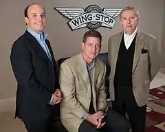 Trois hommes en costumes, dont celui du milieu assis, pose devant un logo blanc Wingstop sur le mur beige.