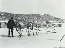 Photo en noir et blanc d'un squelette métallique d'avion posé sur la neige, une personne postant devant.