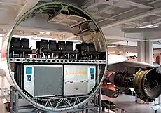 Segment circulaire de fuselage, découpé, en exposition. Sur le plancher de cabine, des rangées de huit sièges, sous celui-ci, deux conteneurs.