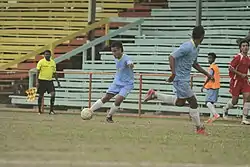 Photographie d'une scène d'un match de football avec un joueur au maillot bleu ciel, faisant une passe à un coéquipier.