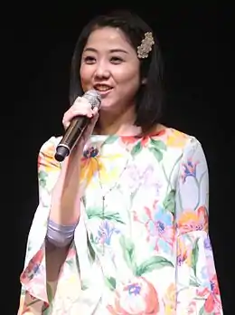 Photographie d'une femme brune d'origine asiatique tenant un micro à sa bouche, porte une robe imprimée de fleurs.