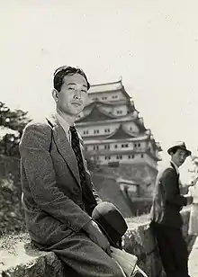 Photographie en noir et blanc d’un jeune homme japonais en costume au premier plan devant un autre homme quelques mètres derrière lui. Un château se trouve à l’arrière-plan.