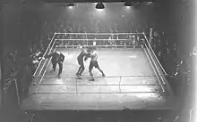 Photographie en noir et blanc d'un ring éclairé sur lequel deux boxeurs s'affrontent sous le regard de l'arbitre et d'une foule de spectateurs.