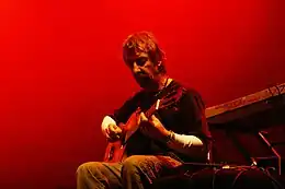 Sur un fonds de lumière rouge, un homme barbu, assis, joue de la guitare.