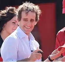 Photographie d'un homme, en chemise blanche, souriant, en plan poitrine, signant un autographe.