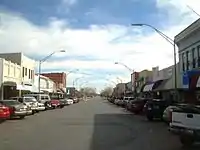 Photographie d'une ville avec une route au centre et des voitures stationnées en épi devant des commerces des deux côtés.