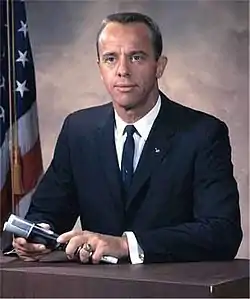 Assis à un bureau, Alan Shepard est habillé d'un veston sombre et tient un microphone dans ses mains. Il porte une bague de la classe de l'Académie navale à sa main gauche et un pin d'astronaute sur le revers du veston. Un drapeau des États-Unis se trouve à l'arrière-plan.Alan Shepard dans les années 1960.