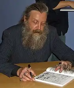 photo d'Alan Moore assis à une table, dédicaçant un album avec un feutre noir, costume sombre, longue barbe grisonnante, bagues métalliques exubérantes à chaque doigts