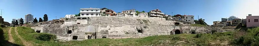 Vue panoramique des vestiges d'un amphithéâtre antiques ; bâtiments modernes en arrière-plan.