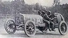 Photographie en noir et blanc d'une automobile numérotée 13A sur laquelle se tient deux hommes, l'un au volant, l'autre assis.