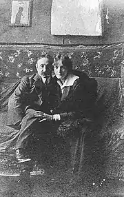 Photographie en noir et blanc représentant assis tout proches l'un de l'autre sur un canapé un homme à tête ronde et à lunettes et une jeune femme blonde en robe longue