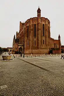 Photo couleur d'une place pavée grise piétonne devant une cathédrale en briques rouges.