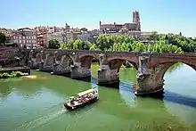 Photo couleur d'une gabarre remplie de touristes. La rivière est verte et la ville en arrière plan en bruques rouges.