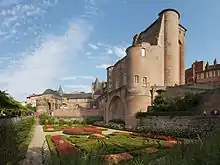 Photo couleur d'un château en brique rouge derrière un jardin à la française. Le donjon se présente comme une tour carrée à quatre tourelles d'angle, dont les deux de gauche ont été raccourcies. La trace des voûtes en ogive sur le mur de droite restant, donne une idée de la grandeur de la bâtisse.