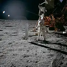 Photo d'un astronaute sur la Lune devant un LEM avec sur sa gauche un collecteur de vent solaire.