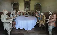 Photographie en couleurs. Portrait de groupe des treize statues très colorées représentant Jésus et ses apôtres autour d'une table sur laquelle on a dressé une nappe blanche.