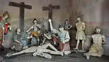 Photographie en couleurs. Alors que deux croix sont peintes sur le mur, un groupe de statutes colorées représente Jésus allongé sur sa croix, au sol, pendant qu'un soldat plante un clou dans sa main, devant des soldats, un dignitaire romain et deux autres condamnés.