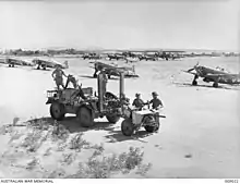 Des soldats, et leur camion tractant un canon antichar, devant des avions de chasse garés sur un terrain d'aviation.