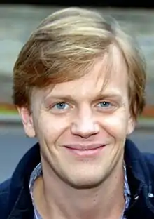 Gros plan d'un homme aux yeux bleus et aux cheveux blonds coiffés avec une raie sur sa gauche. Il regarde le photographe et sourit avec la bouche fermée.