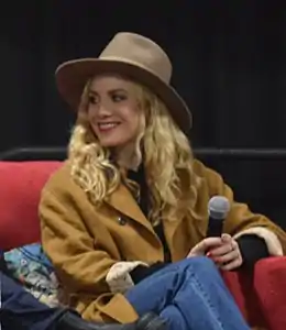 A 34-year-old blonde-haired woman wearing a hat and holding a microphone, smiling to the left of the camera.