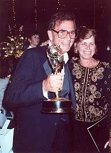 un homme en costume bleu et portant des lunettes soulevant un trophée, à ses côtés une femme portant un haut brodé
