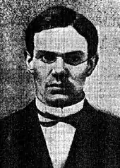 Portrait photographique en noir et blanc d'un jeune homme aux cheveux sombres, soigneusement coiffé et rasé, portant un costume et des lunettes.