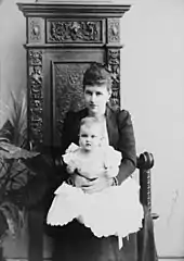 Portrait d'une jeune femme à la robe sombre portant sur ses genoux un bébé à la langue robe blanche.