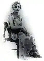 Une jeune femme aux cheveux bouclés et au visage enfantin, vêtue d'une robe à fleurs et assise sur une chaise.