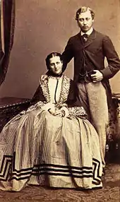 Photographie sépia montrant un couple, elle assisse, lui debout à droite.