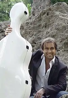 Alexandre Debrus assis devant un rocher tenant de sa main droite la boîte blanche de son violoncelle posée à côté de lui.