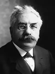 Photographie de face d'un homme portant des lunettes et une moustache.