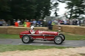 Photographie d'une voiture rouge en piste, le public figurant en arrière-plan derrière des bottes de paille.