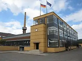 photographie couleur d’une usine prise de trois-quart, façades largement vitrées, grande cheminée industrielle en arrière plan à gauche et drapeaux surplombant l’édifice.