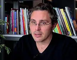 un homme avec de petites lunettes rondes parle devant un rayon de bibliothèque